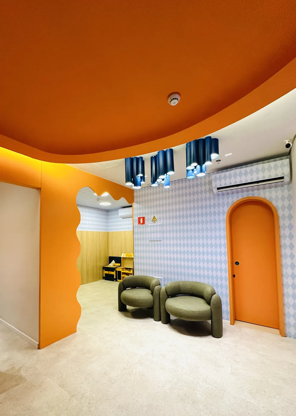Sala de espera com teto laranja ondulado, luminárias azuis e porta em arco